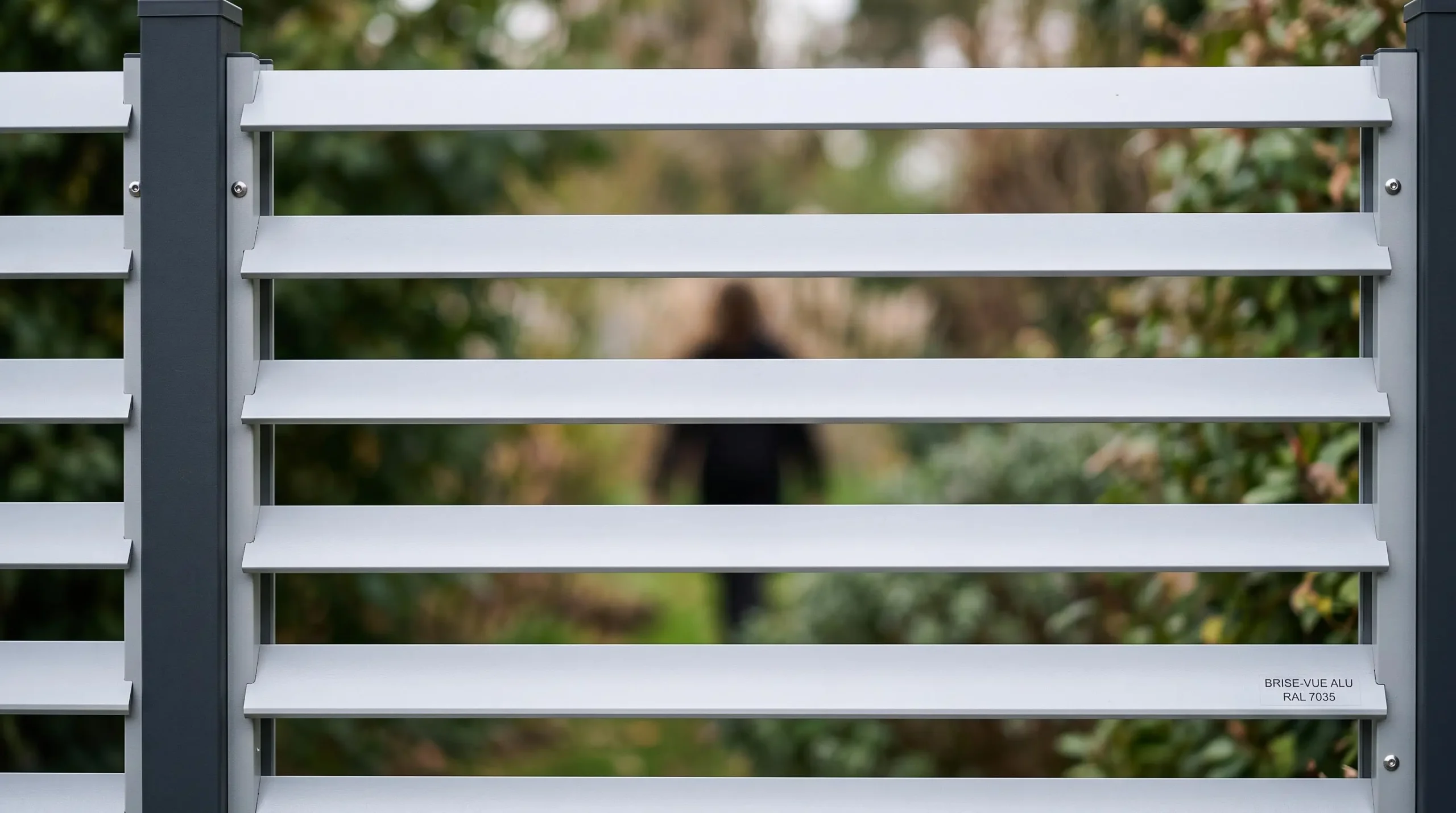 Gros plan sur un panneau de clôture aluminium brise-vue gris clair montrant les lames horizontales espacées qui bloquent la vue tout en laissant passer l'air, silhouette floue visible en arrière-plan